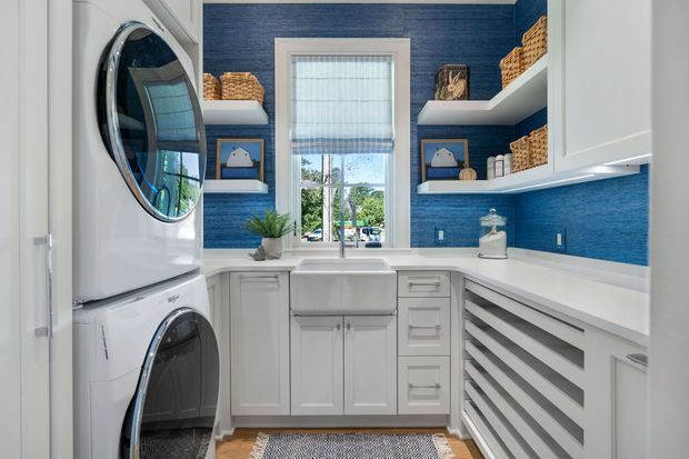 Bright, compact laundry room with white built-in cabinetry, stacked front-loading washer and dryer, a deep farmhouse-style utility sink beneath a window, open shelving with woven baskets, and vivid blue textured wall covering. The room has a clean, organised, high-contrast coastal-inspired fit-out.