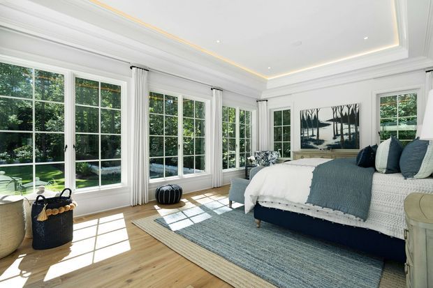 Bright, elegant bedroom or sunroom-style retreat with expansive white-framed multi-pane glazing, layered white wall and ceiling trim, concealed cove lighting, pale timber flooring, and a large upholstered bed dressed in white and blue-grey linens. A textured area rug, woven basket, small ottoman, and monochrome artwork add understated coastal character. Lush greenery is visible outside through the windows.