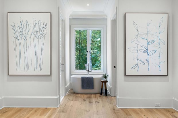 Bright, refined bathroom viewed from a hallway or adjoining room, featuring a freestanding white bathtub positioned beneath a window, pale timber flooring, white walls with decorative lower wall panelling, and matching botanical artworks on either side. A small stool with a potted plant and a dark towel add restrained accents. The room has a calm, light-filled contemporary-traditional feel.