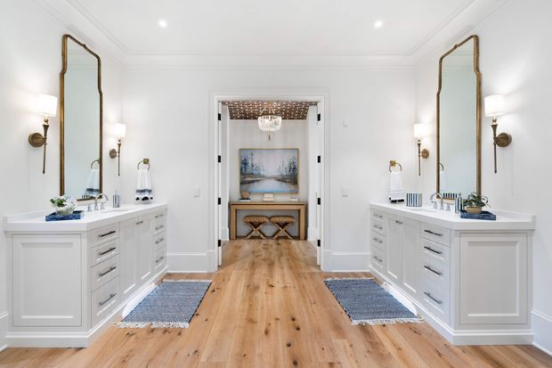 Bright, symmetrical bathroom with two white built-in vanities facing each other, large brass-framed mirrors, wall sconces, and pale timber flooring. A central doorway leads to a small styled sitting or dressing area with artwork, a console, and two stools. The room has refined traditional detailing with a clean, luxurious coastal feel.