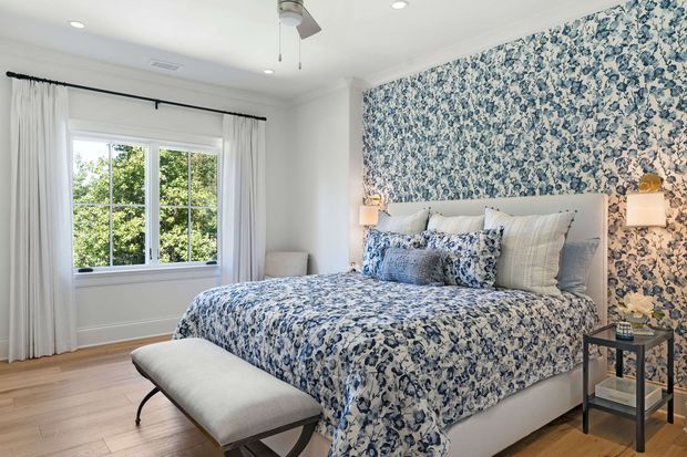 Bright, traditional-style bedroom with a large upholstered bed dressed in blue-and-white floral bedding, matching floral wallpaper covering the feature wall, white curtains, light timber flooring, bedside sconces, and a small upholstered bench at the foot of the bed. A window provides a leafy outlook, while a ceiling fan and recessed lighting complete the room.