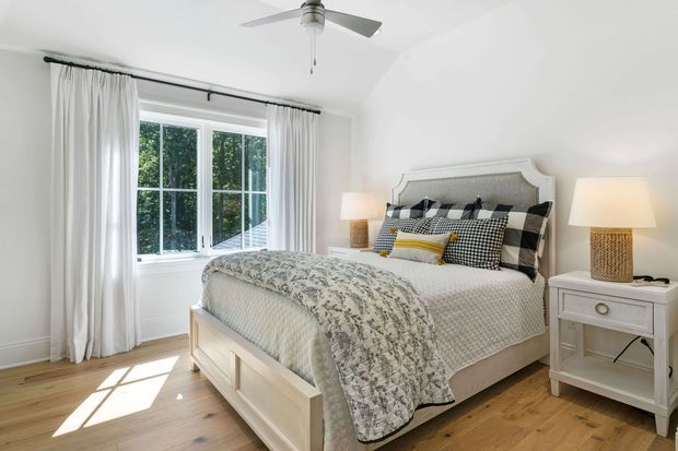 Bright, comfortable bedroom with a white upholstered bed and headboard, layered patterned bedding, matching white bedside tables, woven-texture lamps, sheer white curtains, and a large multi-pane window. The room has light timber flooring, white walls and ceiling, and a ceiling fan, creating a clean, airy, traditional-contemporary interior.