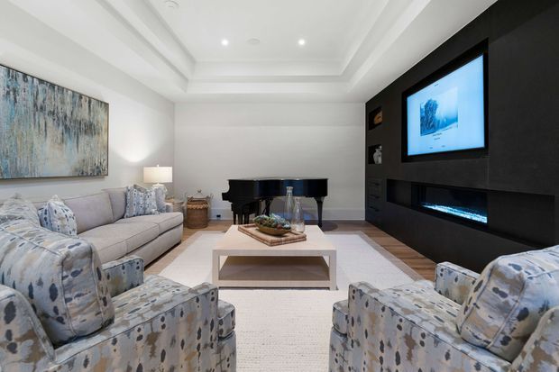 A refined living room with a large sectional sofa and two patterned armchairs arranged around a low rectangular coffee table. The room features a black built-in media wall with television, recessed electric fireplace, and display niches, plus a black grand piano, pale rug, abstract artwork, recessed ceiling detailing, and soft neutral furnishings.