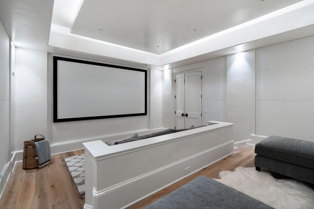 A bright, refined media room with a large wall-mounted projection screen, concealed or built-in cabinetry, recessed cove lighting, and a raised upholstered seating platform or partition. The room features pale timber flooring, white panelled walls and ceiling, black screen framing, and grey upholstered furniture over a light rug.