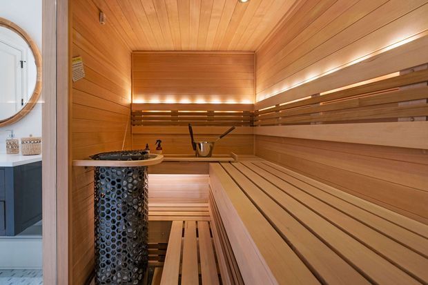 A compact, warmly lit timber sauna viewed from the entry, with extensive horizontal wood wall and ceiling lining, tiered slatted bench seating, integrated linear lighting, and a dark cylindrical sauna heater with stones and a bucket nearby. A bathroom vanity area is partially visible through the doorway at left.