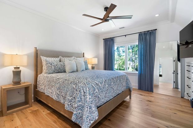 Bright, well-appointed bedroom with a wood-framed bed dressed in a blue-and-white patterned quilt and coordinating pillows. Matching bedside tables and lamps flank the bed, while blue curtains frame a window overlooking greenery. The room has white walls and ceiling, warm timber flooring, recessed lighting, and a contemporary ceiling fan. A mirrored wardrobe or dressing area is visible at right.
