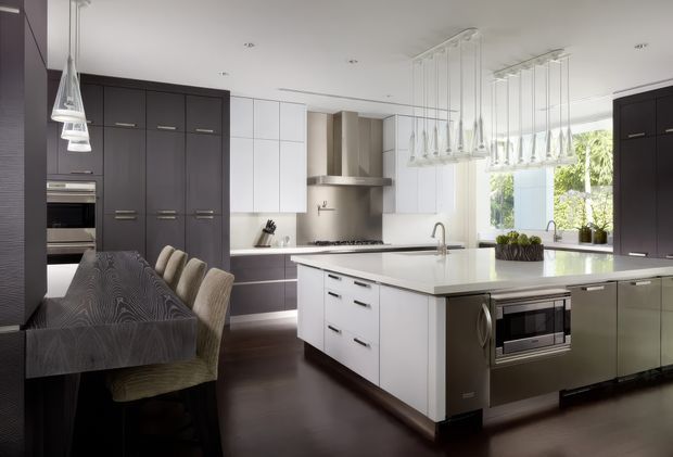 A spacious contemporary kitchen and dining area with dark timber cabinetry, white upper cabinets, a large white island with integrated cooktop and sink, built-in appliances, and a dark timber dining table with upholstered chairs. Multiple sculptural pendant lights hang above the island and dining area, while a large window brings in natural light and greenery views.