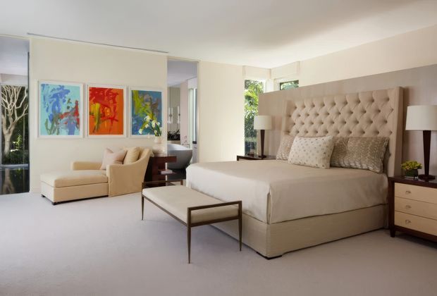 Spacious, softly lit contemporary bedroom with a large upholstered bed featuring a tufted headboard, matching bedside tables and lamps, a bench at the foot of the bed, and a separate lounge area with a chaise and armchair. Pale carpeting, warm neutral walls, and colourful abstract artwork create a refined, comfortable interior.