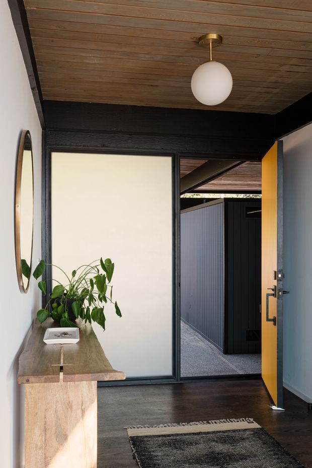 A compact, refined entryway with dark timber flooring, a timber-lined ceiling, black-painted structural framing, and a pale blue-grey wall. A sculptural timber console with a stone-like top sits beneath an oval mirror, accompanied by a leafy plant. A large frosted-glass panel brings diffused light into the hall, while an open mustard-yellow door leads to a covered exterior walkway or courtyard. A simple globe pendant provides overhead lighting.