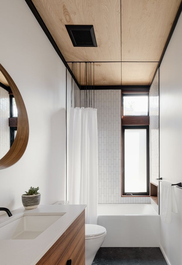 Bright, narrow contemporary bathroom with a white tiled bath/shower enclosure, freestanding white bathtub, toilet, and floating timber vanity with integrated white basin. A large round timber-framed mirror, black tapware and hardware, white curtain, dark floor, and pale timber ceiling create a warm modern interior. A tall window brings in natural light.