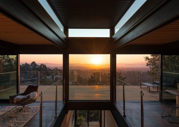 A dramatic contemporary interior opening onto a balcony or terrace, with expansive floor-to-ceiling glazing framing a sunset city and valley panorama. Dark timber ceiling slats and substantial exposed structural beams create a strong central axis around the glazed opening and stair void. The space includes an upholstered lounge chair, outdoor seating, glass balustrades, and warm reflected sunlight.