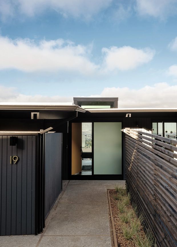 A narrow covered entry walkway leading to a dark, contemporary building entrance. The facade features black vertical cladding, a timber slatted privacy fence, concrete paving, frosted and clear glazing, and a flat roof with a raised clerestory-like element. A house number plaque is mounted on the left gate, with sky visible above the roofline.