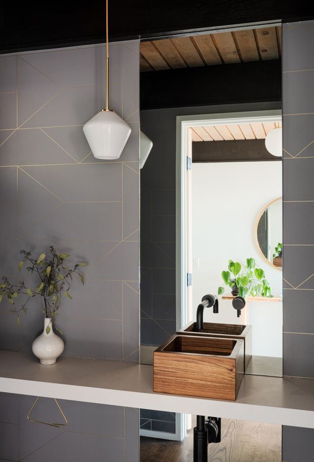 A refined bathroom vanity area with a dark grey geometric tiled wall featuring thin brass-coloured linear detailing. A white pendant light hangs above the stone-look countertop, which holds a timber vessel sink with a matte black tap. A large mirror reflects an adjacent bright space with greenery and a round wall mirror. The ceiling has exposed timber boards and dark structural beams, creating a sophisticated, moody interior with warm natural accents.
