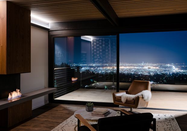A sophisticated living room with expansive floor-to-ceiling glazing overlooking a brightly lit cityscape at night. The room features dark timber built-in cabinetry, a linear fireplace, a slatted timber ceiling with a substantial dark structural beam, warm timber flooring, a sculptural lounge chair with a sheepskin throw, and a low coffee table with a sofa. The interior has a dramatic, refined atmosphere with strong indoor-outdoor visual connection.