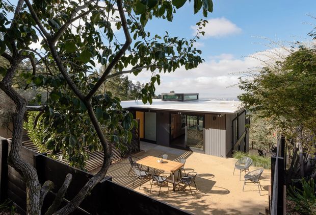Exterior view of a compact modern home set among mature trees, with a flat roof, dark and light vertical cladding, large glazed openings, and a broad outdoor terrace furnished with a dining table and chairs. The elevated deck and surrounding vegetation create a private, leafy setting.