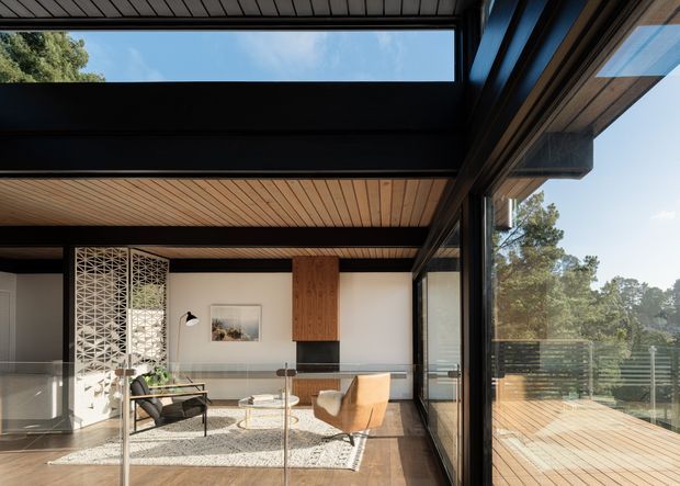 A refined mid-century-inspired living room with floor-to-ceiling glazing, a glazed deck, exposed dark structural beams, and a warm timber-lined ceiling. The room includes a neutral sofa, sculptural lounge chair, low round coffee table, patterned rug, fireplace or dark feature joinery, and a decorative breeze-block screen. Abundant daylight and surrounding trees create a strong indoor-outdoor connection.