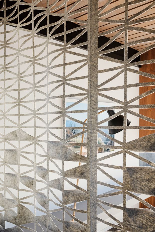 Close-up interior view of a sculptural grey perforated metal or textured screen forming a geometric geometric-patterned stair or balustrade enclosure. Behind it are white walls, warm timber ceiling and wall cladding, and glimpses of a contemporary interior with artwork and a black pendant light. The screen dominates the frame and creates a layered, semi-transparent architectural feature.