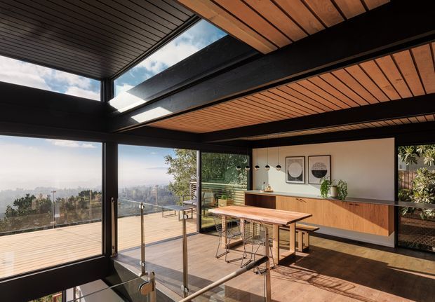 A bright, modern open-plan kitchen and dining area with expansive floor-to-ceiling glazing opening onto a timber deck and distant hillside views. The space features a long timber dining table with stools, a streamlined timber kitchen island and cabinetry, black steel structural framing, warm timber slatted ceilings, polished dark flooring, and glass balustrading around a stair opening. Strong indoor-outdoor connection and abundant natural light define the scene.