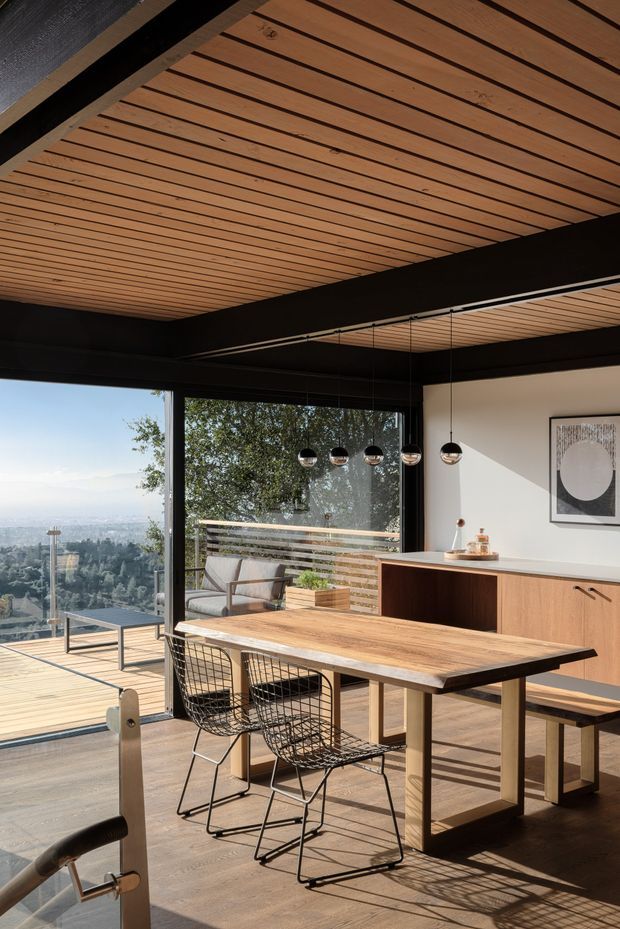 A warm, modern dining and kitchen space with a large timber dining table, black wire-frame chairs, and a timber kitchen island with a pale benchtop. Full-height black-framed glazing opens to a timber deck and expansive landscaped city view. The ceiling features repeated timber slats between bold dark structural beams, with a row of spherical pendant lights above the island. The room has abundant natural light and a refined indoor-outdoor connection.
