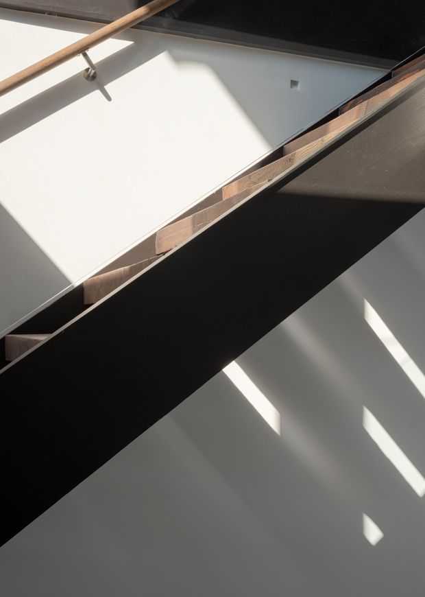 Abstract architectural interior view looking upward at a stairway, with a dark solid stair stringer or balustrade crossing diagonally through the frame, exposed timber stair treads, and a timber handrail mounted against white walls. Strong sunlight creates geometric shadows across the walls, emphasizing the sculptural form and high-contrast composition.