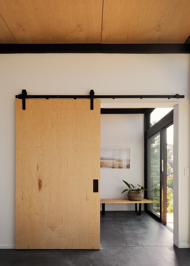 Interior view of a contemporary transitional space featuring a large natural timber sliding barn door on exposed black metal hardware. The door opens toward a bright sitting or entry area with a built-in timber bench, black-framed floor-to-ceiling glazing, dark polished concrete flooring, white walls, and a timber-lined ceiling. A potted plant and framed artwork add understated decoration.