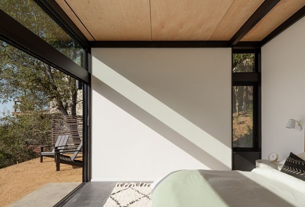 A compact, light-filled bedroom with floor-to-ceiling black-framed glazing opening toward a natural outdoor setting. The room has a pale timber panel ceiling with exposed dark structural beams, smooth white walls, a dark floor, and a partially visible bed with pale green bedding. Strong diagonal sunlight creates a graphic shadow across the wall, while a simple wall light and bedside table provide restrained detailing. An outdoor chair is visible beyond the glazing.