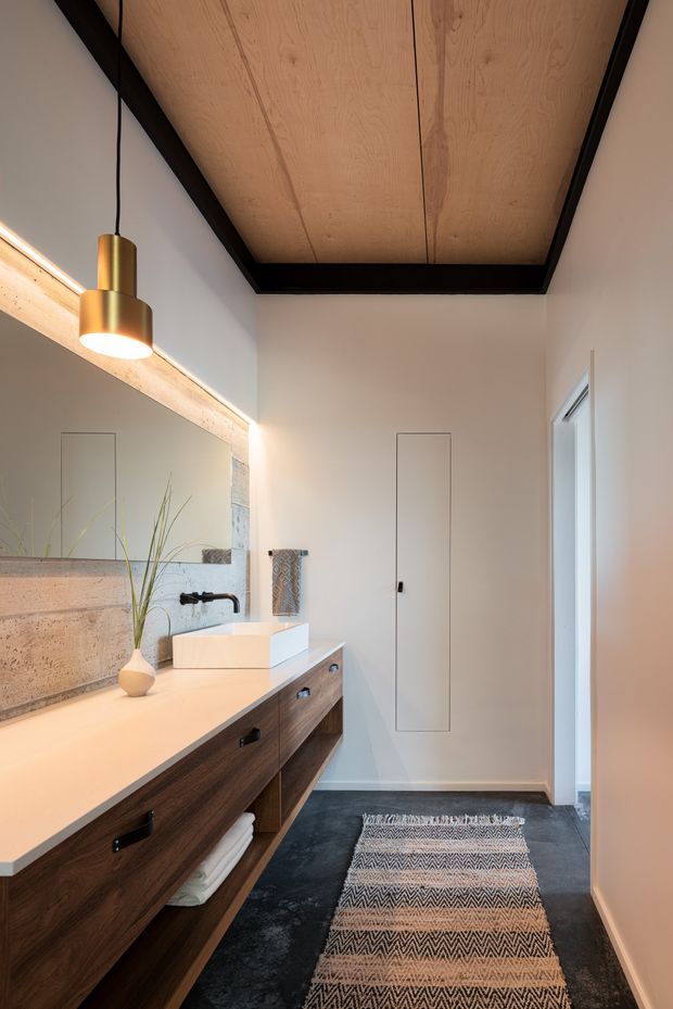 A narrow, refined bathroom featuring a long floating dark timber vanity with open shelving, a pale stone or solid-surface benchtop, and a white vessel basin. A large illuminated mirror runs along the left wall above a textured concrete or stone backsplash. The room has a dark floor, white walls, a timber-panel ceiling framed with exposed dark beams, a black wall-mounted tap, concealed storage, and a warm cylindrical pendant light. A patterned runner adds softness along the floor.