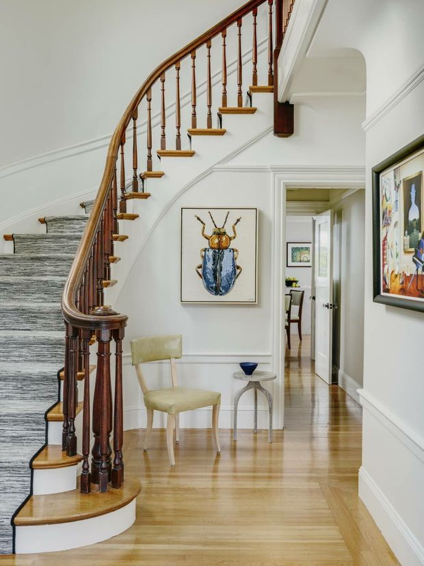 Elegant residential stair hall with a curved staircase, polished dark timber handrail and turned balusters, carpeted treads, white paneled walls, and light timber flooring. A small upholstered chair and side table sit beneath framed artwork, while a doorway leads toward another room. The space has refined traditional detailing and abundant natural light.