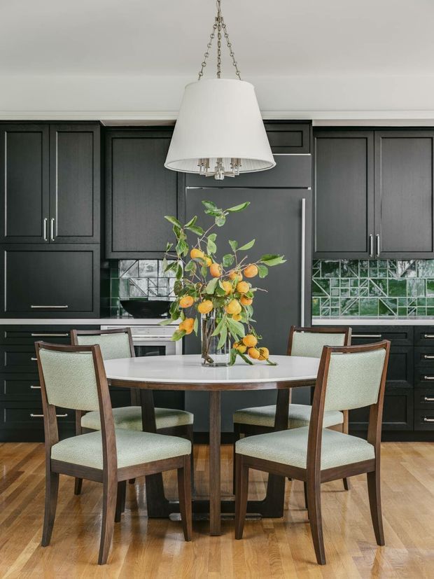 A sophisticated kitchen-dining area with dark charcoal built-in cabinetry, a matching integrated refrigerator, and a glossy green geometric tile splashback. A round white-topped dining table with six upholstered wooden chairs occupies the foreground beneath a large white pendant light. Light timber flooring, stainless hardware, and a leafy arrangement with oranges add contrast and warmth.