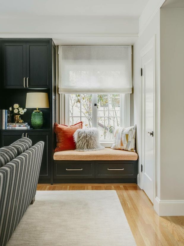 A refined living room corner featuring a built-in dark painted cabinetry wall with upper cupboards, open shelving, and lower drawers surrounding a cushioned window seat. The window has a white Roman blind, while patterned and textured cushions add warmth. Light timber flooring, white walls and trim, a striped upholstered armchair, and a pale area rug create a classic, inviting interior.