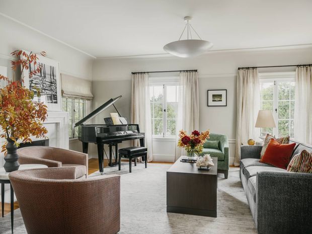 A bright, refined living room with tall multi-pane windows dressed in sheer curtains, a black grand piano, two rounded upholstered armchairs, a green accent chair, a grey sofa with patterned and rust cushions, and a dark rectangular coffee table. The room has light neutral walls, a pale carpet, decorative fireplace or mantel detailing, framed artwork, a sculptural ceiling pendant, and floral arrangements. The overall atmosphere is elegant, comfortable, and classically styled.