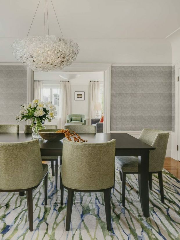 Elegant dining room with a large black rectangular dining table, upholstered green-grey dining chairs, patterned area rug, and a sculptural crystal chandelier. Textured grey wall coverings with white trim frame an open connection to a softly furnished living room, while polished timber flooring is visible at the edges. The room has a refined, layered, contemporary-traditional character with abundant natural light.