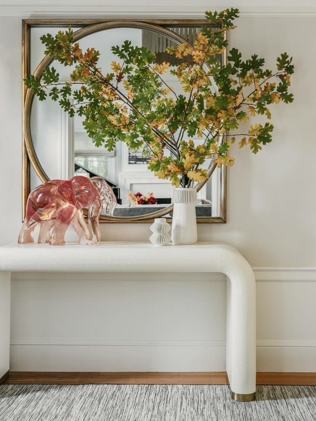 A refined interior vignette featuring a rounded white console table against a paneled light wall, topped with a white ceramic vase of leafy branches, a small sculptural vase, and a translucent pink glass elephant. A large antique-style arched mirror with a metallic frame hangs above the console, reflecting an elegant room beyond. Dark timber flooring and a grey patterned rug add contrast.