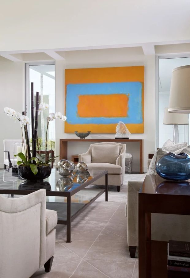 Bright, refined living room with cream upholstered armchairs and sofa, a dark wood and glass coffee table, pale patterned rug, and a dark wood console. A large abstract orange-and-blue artwork forms the focal point above the console, while orchids, metallic sculptures, and blue glass decor add accents. Full-height windows and a contemporary coffered ceiling bring in abundant natural light.
