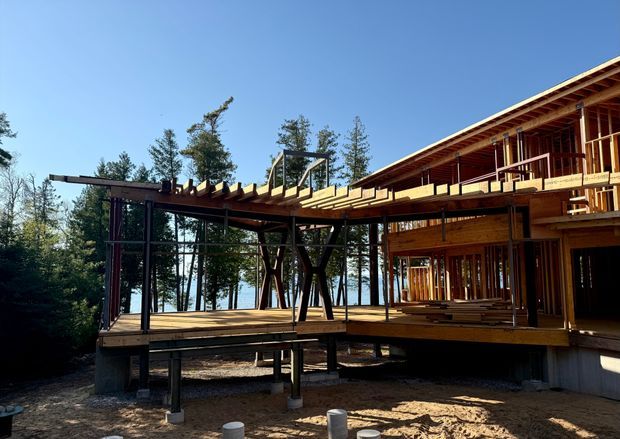 Exterior view of a timber-framed multi-level house under construction in a wooded setting, with extensive exposed structural posts, beams, rafters, raised decks, and open glazing areas. The building sits elevated above a rough sandy construction site, with a lake or bright water visible through the trees in the background.