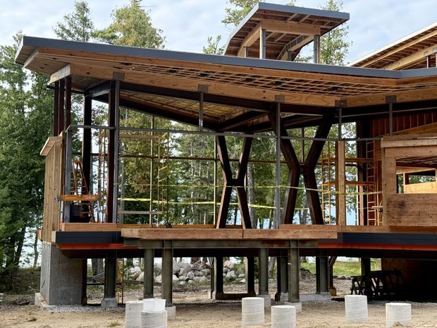 Exterior view of a partially constructed, elevated timber-and-steel building set among trees. The structure features extensive floor-to-ceiling glazing, exposed timber roof framing, dark structural posts and beams, cantilevered roof planes, and concrete pier foundations. A raised roof platform or clerestory-like pavilion is visible above the main roofline.