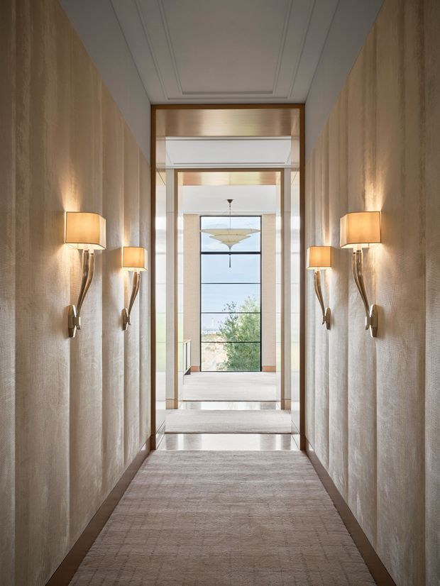 A refined, narrow residential hallway with textured taupe walls, symmetrical sculptural wall sconces, a pale carpet runner, dark timber skirting, and a coffered white ceiling. The corridor opens toward a bright landing with large black-framed glazing and warm timber detailing, creating a layered, axial perspective.