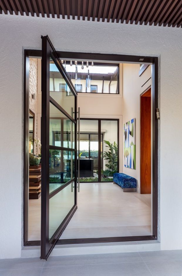 A contemporary interior view through a large dark-framed glazed pivot door into a bright double-height entry hall or courtyard-like foyer. The space features pale flooring and walls, extensive internal glazing, a sculptural hanging light, a blue upholstered bench, indoor planting, colourful artwork, and a dark timber slatted ceiling visible in the foreground. A stair and additional rooms are visible through the glass.