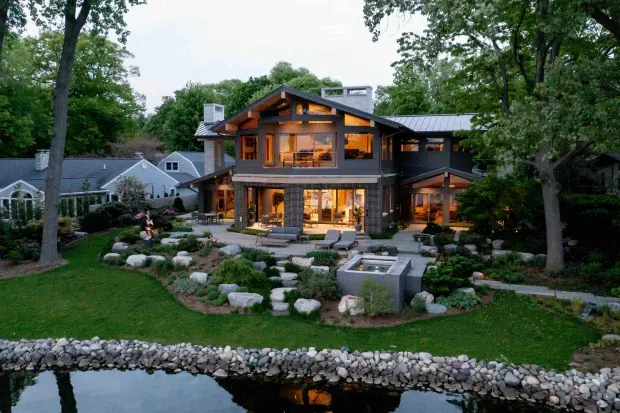 Exterior view of a large multi-level contemporary home set in a heavily landscaped suburban garden beside a narrow water feature. The house has dark masonry and timber elements, extensive glazing, prominent exposed gable framing, and a standing-seam metal roof. Warm interior lighting glows through the windows and doors, with terraces, outdoor seating, stone landscaping, and a small square water feature visible in the foreground.