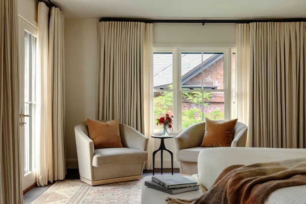A refined bedroom sitting area with two upholstered armchairs positioned beside a window, framed by full-height neutral drapery. A small side table, layered rug, coffee table, books, and a partially visible bed with a throw create a warm, elegant retreat. The room has traditional detailing, balanced natural light, and a muted cream, beige, and caramel palette.