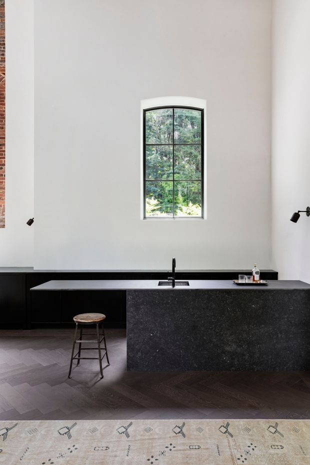 Minimalist kitchen interior with a long black island and matching dark cabinetry, a pale stone benchtop, black undermount sink and tap, and a tall arched multi-pane window overlooking greenery. The room has high white walls, small black wall lights, dark herringbone timber flooring, and a patterned rug in the foreground.