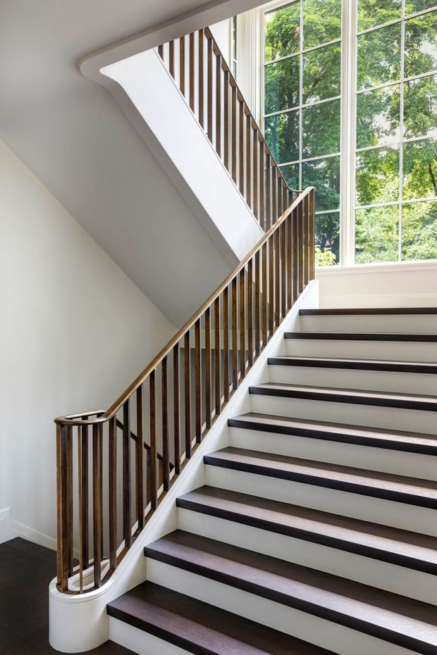 Bright interior view of a refined stairway with dark timber stair treads, white painted risers and walls, and a curved timber balustrade with slender vertical spindles. A large divided-light window brings in abundant daylight and overlooks leafy greenery. The stair design combines clean contemporary detailing with traditional proportions and trim.