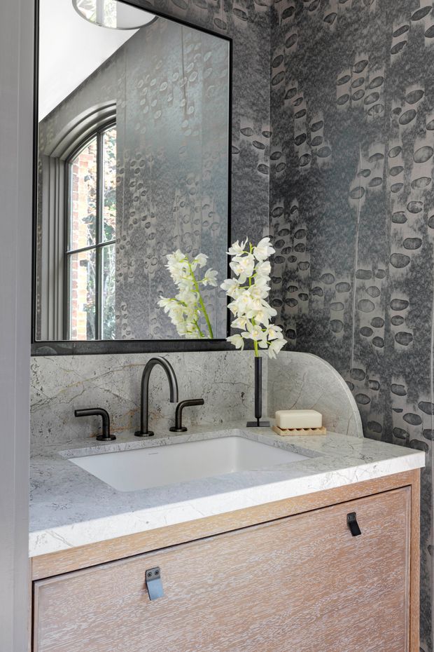 A refined bathroom vanity featuring a pale stone countertop and integrated rectangular basin, dark matte metal wall-mounted-style tapware, and light timber drawer cabinetry. The backsplash and surrounding walls are clad in dramatic dark grey patterned stone or tile with organic oval markings. A large black-framed mirror reflects an arched window, while white flowers provide a small decorative accent.