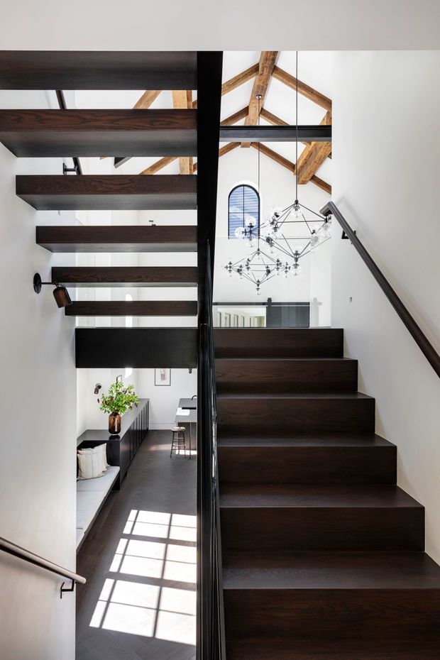 A modern interior stairway with dark timber treads and matching open-riser staircase elements, black metal balustrade, and white walls. The stairwell rises through a bright double-height space with exposed timber roof trusses, a large sculptural geometric chandelier, and glimpses of a minimalist living area below.