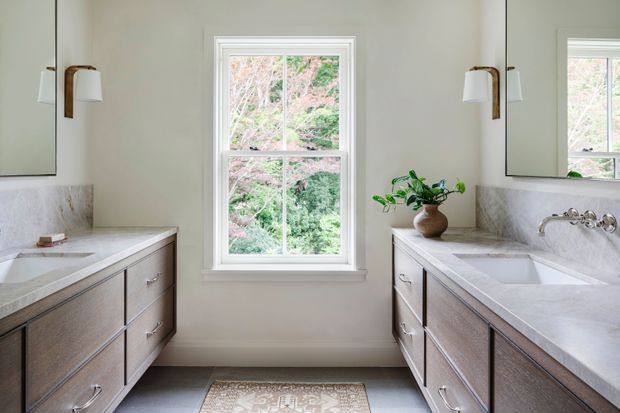 Bright, symmetrical bathroom with twin floating wood vanities, white marble-look countertops and splashbacks, undermount sinks, wall-mounted fixtures, large mirrors, brass wall sconces, and a central double-hung window overlooking greenery. The space has a refined, calm, traditional-contemporary character with abundant natural light.