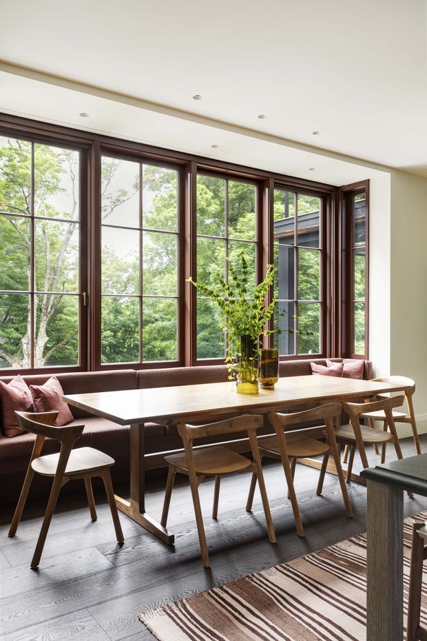 A refined dining area with a long timber dining table surrounded by sculptural wooden chairs and a built-in upholstered banquette. Large dark timber-framed grid windows fill the wall, overlooking leafy greenery and bringing in abundant natural light. The room has pale walls, recessed ceiling lights, dark timber flooring, and a striped neutral rug.