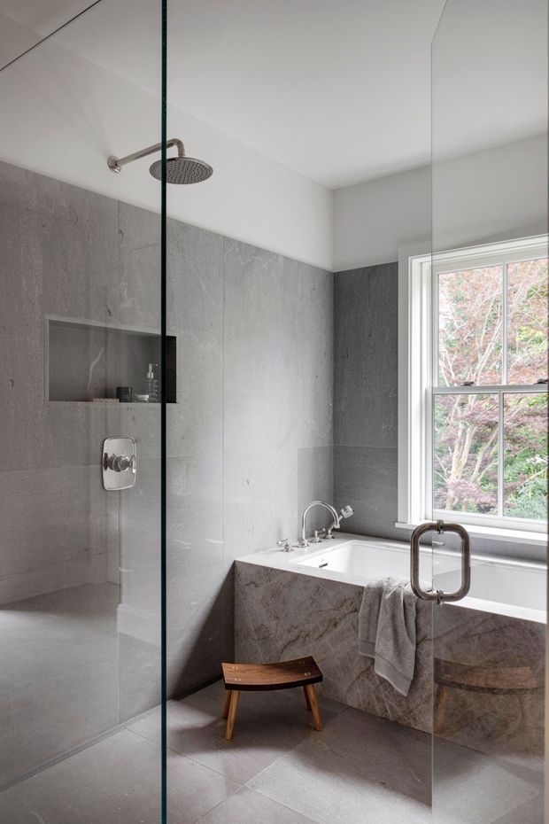 A refined modern bathroom featuring a frameless glass walk-in shower with a ceiling-mounted rain shower, large grey stone-look wall and floor tiles, and a freestanding-style rectangular bathtub clad in matching stone. A large window brings in natural light, while brushed metal plumbing fixtures, a small wooden stool, and a neutral towel add understated warmth.