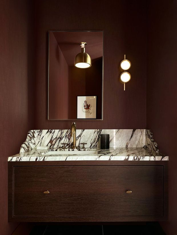 A richly detailed bathroom vanity against deep burgundy-brown wall panelling. The floating dark timber vanity has a white marble countertop and integrated backsplash with dramatic dark veining, a brass-toned faucet and hardware, and a rectangular mirror. Warm brass wall lights and a reflected pendant create an intimate, moody, luxurious atmosphere.
