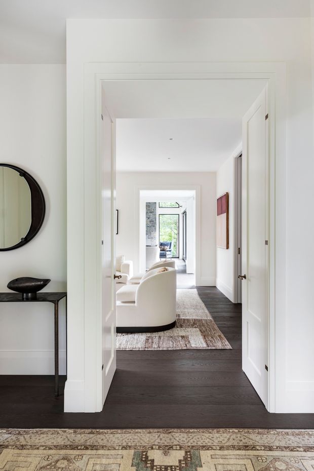 Bright, refined interior view through a wide white-trimmed doorway into a hallway and sitting area. The space features dark timber flooring, white paneled doors and walls, a curved cream upholstered armchair, a slim dark console table with sculptural decor, a round wall mirror, framed artwork, and layered patterned rugs. Daylight enters from glazed doors or windows at the far end, creating a calm, elegant residential atmosphere.