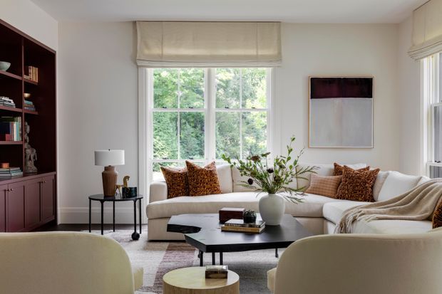 A refined living room with a large multi-pane window overlooking greenery, a pale sectional sofa, two upholstered lounge chairs, a black low-profile coffee table, and a built-in dark wood bookcase with lower cabinetry. Neutral walls and furnishings are accented by patterned rust-toned cushions, a soft throw, abstract wall art, and organic floral branches.